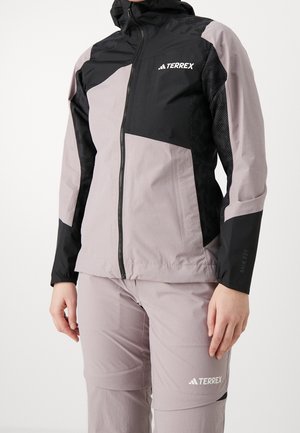 Personne portant une veste imperméable Adidas Terrex gris clair et noir ainsi que un pantalon assorti, debout devant un fond uni.