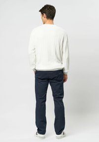 Bărbat văzut din spate, purtând un pulover alb cu model în relief, pantaloni albastru închis și adidași albi cu dungi roșii și albastre.