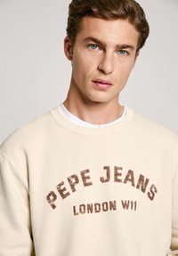 Sudadera beige con cuello redondo y mangas largas, que presenta un gráfico desgastado en rojo que dice "PEPE JEANS LONDON W11". Textura de tela suave.