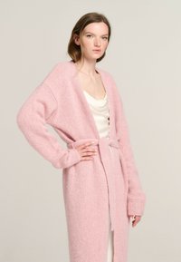 Langer, offener Cardigan aus weichem, rosa Strick. Mit weitem Ärmel und passendem Gürtel. Glatte Textur mit einer gemütlichen Ausstrahlung.