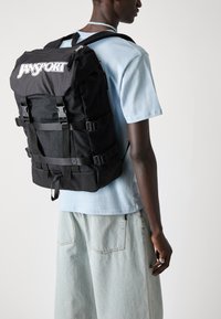 Un sac à dos Jansport noir avec un design texturé, des sangles réglables et des boucles. L'arrière-plan est clair, mettant en valeur les caractéristiques du produit.