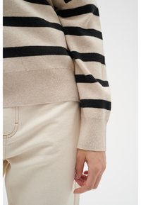 Pull à rayures beige et noir avec poignets côtelés, associé à un pantalon de couleur claire. La texture douce et la coupe décontractée sont évidentes.