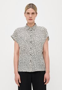 Blouse à manches courtes en crème avec un motif de points irréguliers noirs, présentant une fermeture à boutons sur le devant et un col pointu. Texture de tissu lisse.