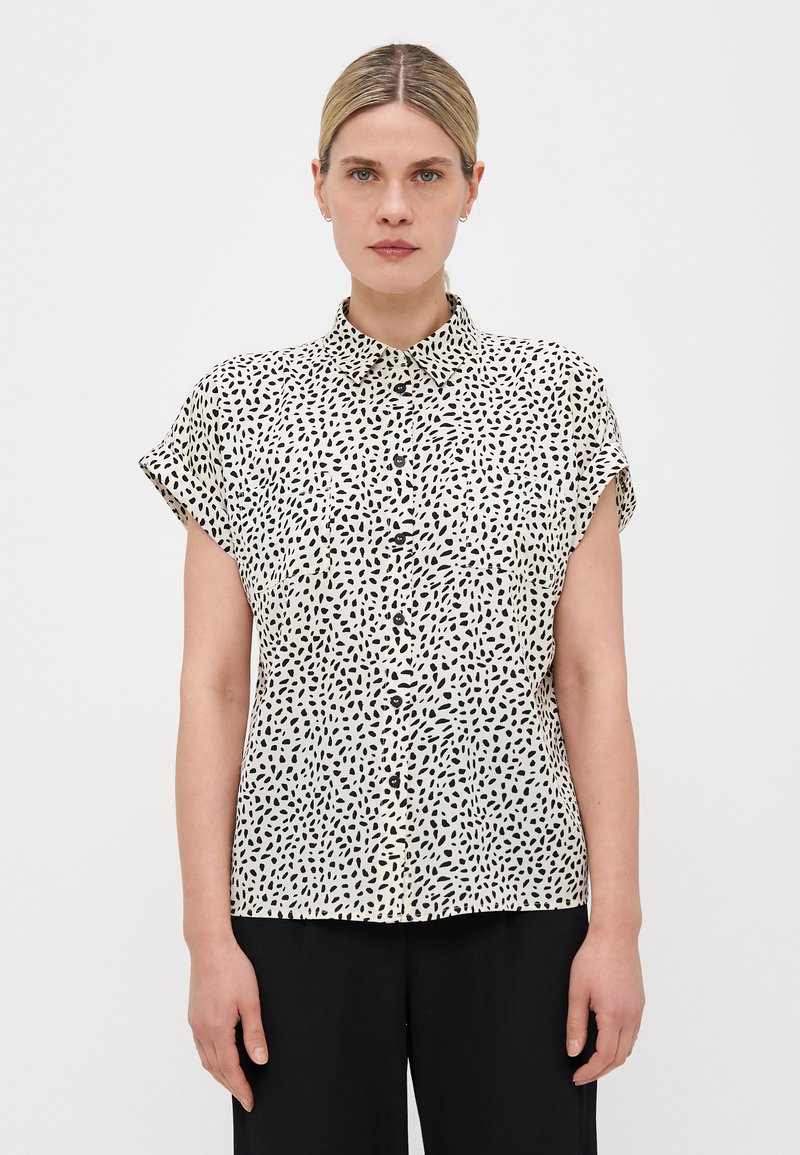Blouse à manches courtes en crème avec un motif de points irréguliers noirs, présentant une fermeture à boutons sur le devant et un col pointu. Texture de tissu lisse.