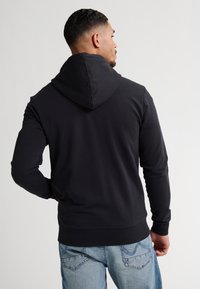 Schwarzer Kapuzenpullover mit langen Ärmeln und geripptem Saum, der eine glatte Textur aufweist. Getragen mit hellblauen Jeans, von hinten gesehen.