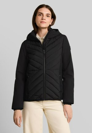 Vrouw draagt een zwart doorgestikt capuchonjack met ritssluiting, gecombineerd met lichtbeige broek, staand tegen een egale grijze achtergrond.