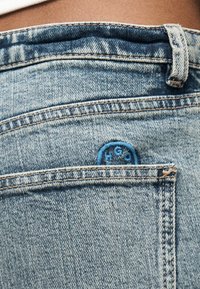 Prim-plan al buzunarului din spate al unei perechi de blugi denim albastru deschis, cu o etichetă circulară mică albastră a mărcii parțial vizibilă în interiorul buzunarului.