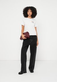 Hvid kortærmet t-shirt med farverig broderi, kombineret med sort højtaljede jeans og en bordeaux clutch. Hun har sorte sko på.