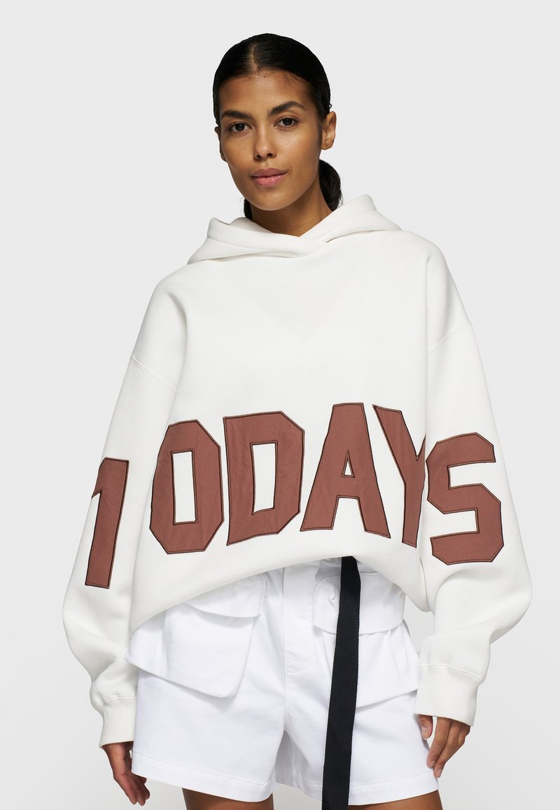 Sweatshirt oversize blanc avec capuche et grand lettrage "10 DAYS" marron en bas. Associé à un short blanc.
