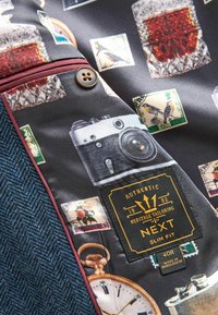 Schlank geschnittenes Jackeninnere mit einem dunklen gemusterten Futter, das Vintage-Objekte zeigt, darunter eine Kamera, eine Uhr und Briefmarken. Blaues Fischgrätenmuster.