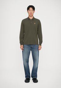 Oliven grønn langermet polo skjorte med krage, med et lite flerfarget emblem; parret med lyseblå rette jeans og svarte sko.
