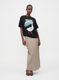 Γυναίκα που φοράει μαύρο μπλουζάκι Moschino με άσπχη χήνα και μπλε ομπρέλα, μπεζ μακριά φούστα με πλαϊνά κουμπιά και μαύρα σανδάλια με τακούνι.