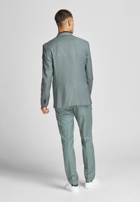 Completo grigio con vestibilità slim e tessuto testurizzato, caratterizzato da uno spacco singolo, abbinato a sneakers bianche. Visto da dietro.