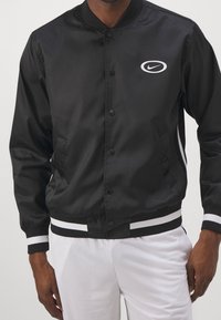 Černá bomber bunda s hladkým povrchem, patentkami, bílými pruhy na manžetách a spodním okraji a bílým logem Nike na hrudi.