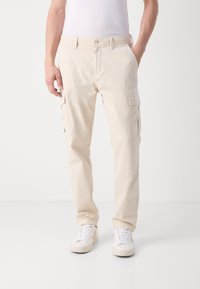 Pantalones cargo beige hechos de un tejido ligero, con bolsillos laterales, cierre de botón y un ajuste relajado, combinados con zapatillas blancas.