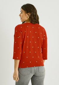 Sudadera roja con mangas tres cuartos, con un patrón de pequeños corazones dorados por todo el tejido. Tela suave y texturizada, ajuste cómodo y escote redondo.