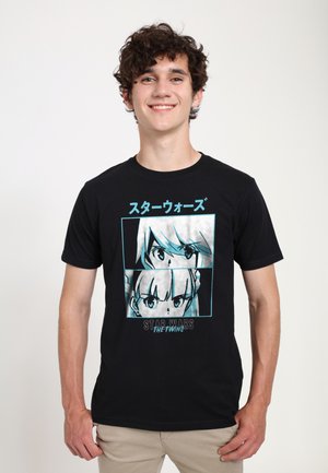 Młody mężczyzna uśmiechający się, ubrany w czarną koszulkę z grafiką oczu w stylu anime oraz japońskim tekstem, stojący na tle jednolitym w jasnym kolorze.