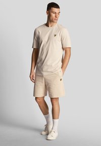 Beige Baumwoll-T-Shirt mit kurzen Ärmeln und einem kleinen gelben Logo auf der Brust, kombiniert mit passenden Shorts und weißen Sneakern.