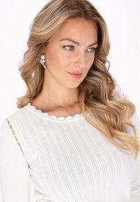 Top alb cu tricot, având o textură ribbed, decolteu în valuri și accente de perle pe mâneci. Modelul are părul ondulat și un machiaj natural.