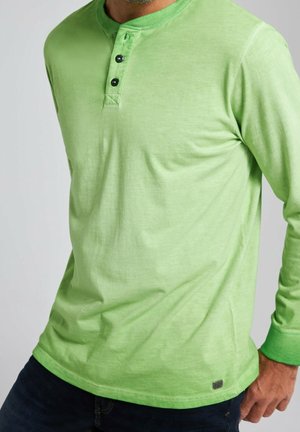 Homme portant un haut henley à manches longues vert clair avec trois boutons foncés et un pantalon foncé, debout devant un fond uni.