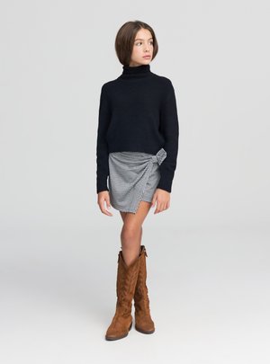 Giovane donna in piedi, con maglione a collo alto nero, minigonna a quadri con dettaglio a nodo e stivali marroni in pelle scamosciata al ginocchio, che guarda di lato.