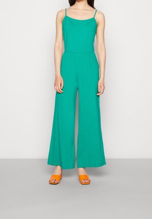 Turkusowy kombinezon na cienkich ramiączkach, luźny krój i szerokie nogawki. W zestawieniu z pomarańczowymi sandałami na paskach. Gładka, lekka tkanina.