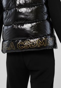 Musta, kiiltävä toppatakki, jossa on uurteinen pinta ja kultainen teksti "OLACSDMNT" helmassa. Yhdistetty mustiin housuihin, mikä korostaa materiaalien välistä kontrastia.