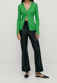 Kvinna iförd en ljusgrön knappad kofta med svarta läderbyxor och svarta spetsiga slingback-sandaler, står framför en neutral bakgrund.