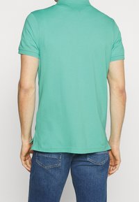 Man som bär en mintgrön kortärmad polo och blå jeans, sedd bakifrån mot en vit bakgrund.