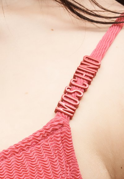 Κοντινή λήψη ενός ροζ υφασμάτινου ιμάντα ρούχου με προσαρτημένα ανάγλυφα γράμματα που γράφουν "MOSCHINO" σε ανοιχτό δέρμα.