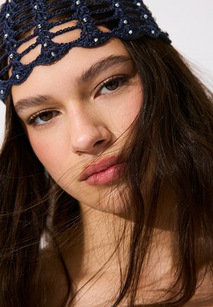 Ung kvinde med langt brunt hår iført et navyblåt hæklet pandebånd med små sølvperler, der blødt kigger på kameraet.