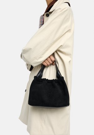 Osoba w beżowym prochowcu trzymająca małą czarną zamszową torebkę z skórzanymi uchwytami blisko ciała.