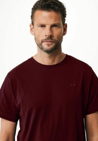 Bordowa koszulka z krótkim rękawem wykonana z miękkiego materiału, z okrągłym dekoltem i małym czarnym logo na lewej piersi.