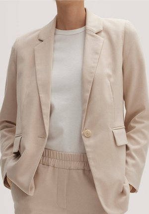 Persoon draagt een beige blazer en bijpassende broek met elastische taille, gecombineerd met een effen witte T-shirt met ronde hals, handen in de zakken.