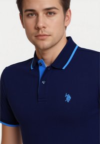 Mladý muž s krátkymi hnedými vlasmi má na sebe námornícky polokošeľu s svetlomodrým golierom a logom, hľadí priamo do kamery proti bielemu pozadiu.