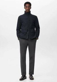 Hombre con una chaqueta negra con cremallera y bolsillos tipo solapa, pantalones grises a medida y zapatos de vestir de cuero negro, de pie contra un fondo liso.