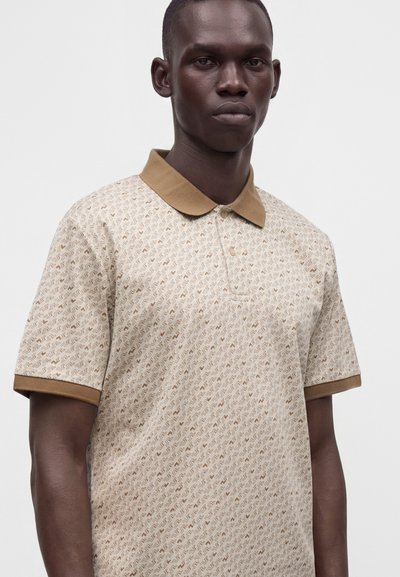 Homme portant un polo beige à motif à manches courtes avec un col et des bords de manches marron, debout devant un fond uni.