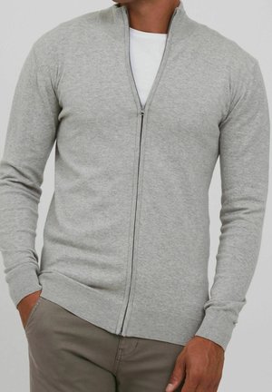 Homme portant un pull à fermeture éclair gris clair par-dessus une chemise blanche, les mains dans les poches, sur un fond neutre.