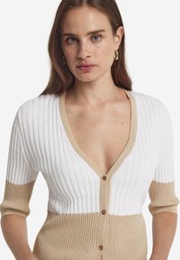Stickad kavaj i vitt och beige, med ribbad textur, djup V-ringning, korta ärmar och dekorativa knappar längs fram.