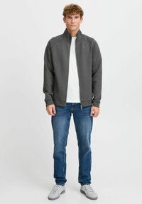 Gray sweatshirt with a high collar, zippered front, and raglan sleeves. Worn over a white T-shirt, paired with blue jeans and gray sneakers.