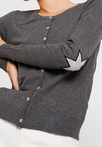 Närbild av en person som bär en mörkgrå knappad cardigan med en vit stjärnpatch på armbågen, tillsammans med vita byxor.