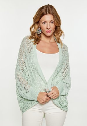 Vrouw met golvend haar die een lichtgroene gebreide sjaal, een witte top en broek en kleurrijke ronde oorbellen draagt, poserend tegen een effen achtergrond.
