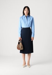 Light blue button-up shirt, navy pleated skirt, beige handbag with pattern, and cream pointed-toe heels. Simple, tailored design elements.