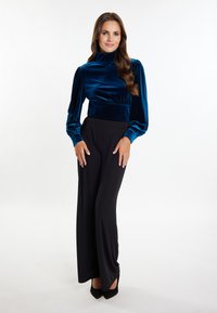 Samtige, türkisfarbene Rollkragenoberteil mit Puffärmeln, kombiniert mit schwarzen, weit geschnittenen Hosen. Schwarze High Heels runden das Outfit ab.