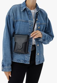 Borsa a tracolla nera in pelle con finitura testurizzata, caratterizzata da una patta pieghevole e logo dorato, indossata sopra una giacca in denim azzurro chiaro.