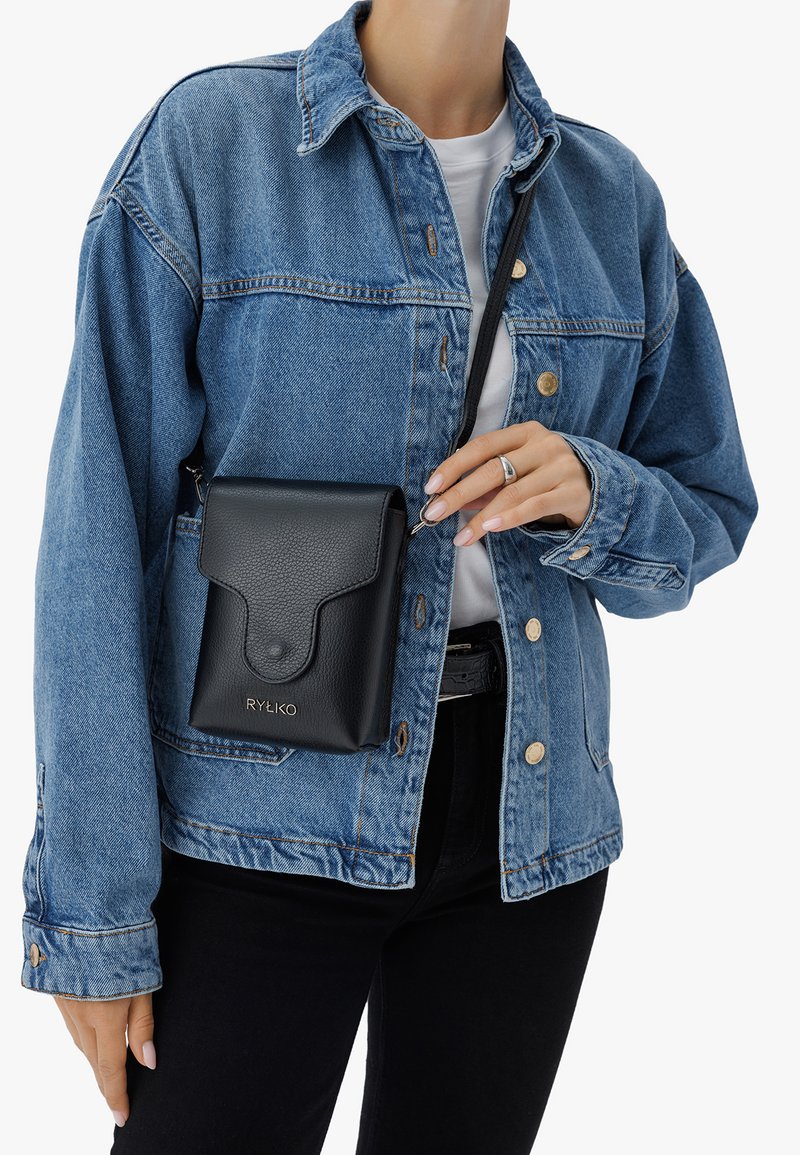 Borsa a tracolla nera in pelle con finitura testurizzata, caratterizzata da una patta pieghevole e logo dorato, indossata sopra una giacca in denim azzurro chiaro.