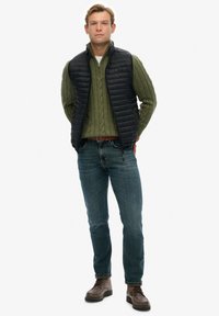 Chaleco acolchado negro sobre un suéter de punto trenzado verde, jeans azules y botas marrones de cordones. Textura suave y detalles de diseño en capas.