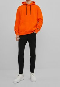 Oranžová mikina s kapucí, přední kapsou a černými šňůrkami, doplněná o černé slim kalhoty a bílé tenisky. Hladký povrch textilie.