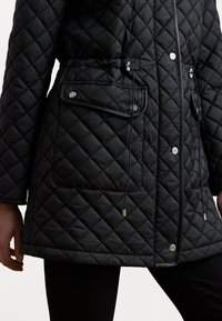 Veste matelassée noire avec taille à cordon, boutons-pressions et poche à rabat. Le matériau a une texture lisse avec des finitions contrastantes.