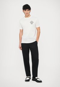 Biała bawełniana koszulka z szarą grafiką w kształcie diamentu, do tego czarne spodnie i czarno-białe mokasyny z złotymi akcentami.